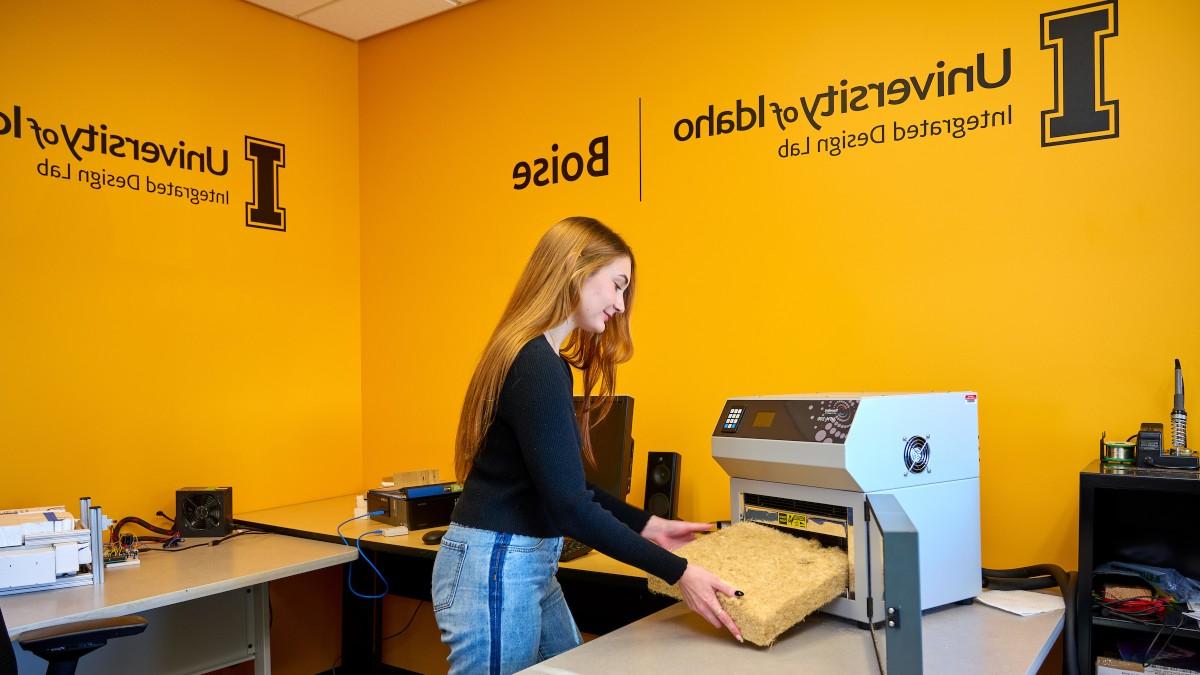 A female student uses equipment in the Integrated Design Lab. The wall behind her is pride gold stenciled with the logo of the University of Idaho Integrated Design Lab and the word Boise. 